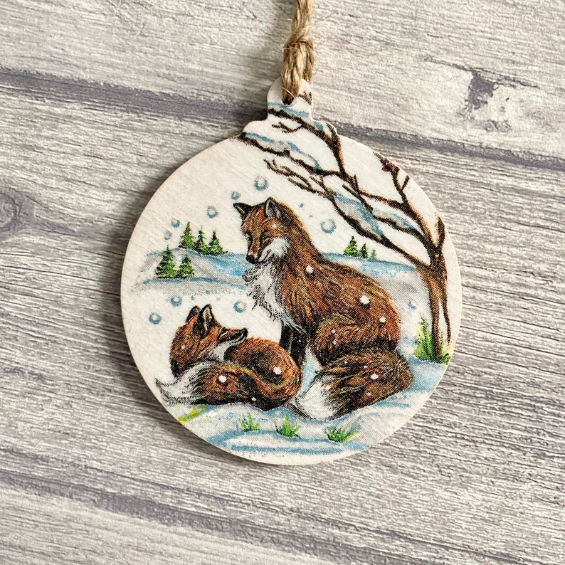May include: A round wooden ornament featuring a printed illustration of two foxes in a snowy landscape. One fox sits upright while the other rests nearby. The scene includes a bare tree, small evergreen trees, and falling snowflakes. A jute twine loop is attached.