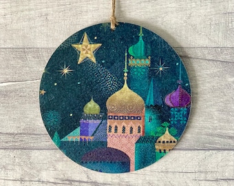 May include: A circular hanging ornament featuring a dark blue night sky with a golden star and a cluster of buildings with colorful domes and spires in shades of gold, purple, teal, and pink, suspended by a thin brown twine string.