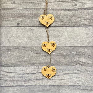 May include: A vertical hanging decoration consisting of three yellow wooden hearts connected by thin twine. Each heart features a repeating pattern of small black and brown bees printed on the surface. The hearts are arranged in a single column.