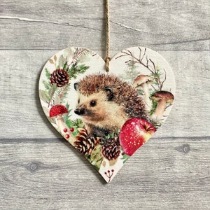 May include: Heart-shaped wooden ornament featuring a detailed illustration of a hedgehog surrounded by pine cones, mushrooms, and a red apple. The ornament is suspended by a natural twine string, perfect for seasonal or woodland-themed decor.