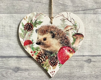 May include: A heart-shaped wooden hanging ornament featuring a detailed illustration of a hedgehog surrounded by pine cones, mushrooms, and a red apple. The design includes green foliage and small red berries, all suspended by a thin twine string.