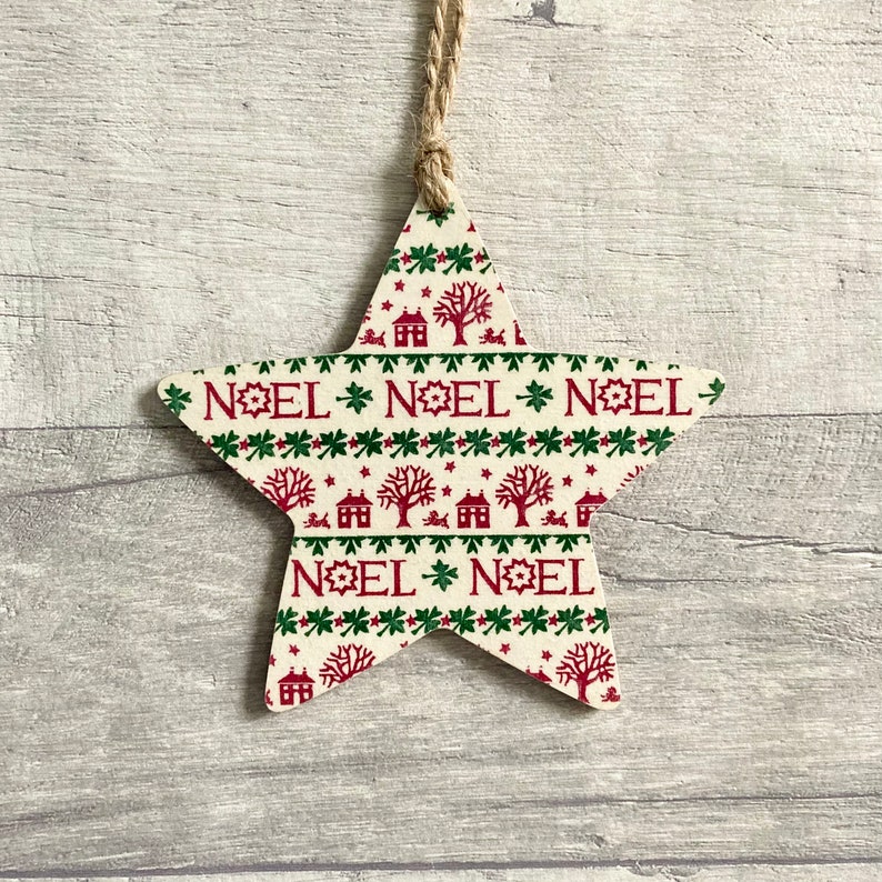May include: A star-shaped wooden ornament featuring a cream-colored background with a repeating pattern of red houses, trees, and the word NOEL in red text. The ornament is suspended by a thin, light brown jute twine loop attached at the top point.