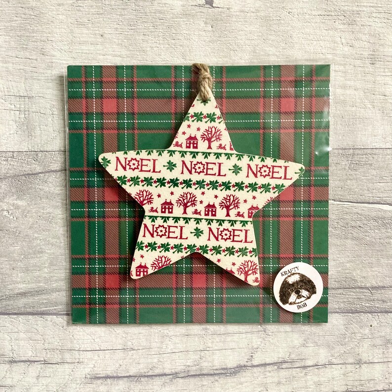 May include: A star-shaped wooden ornament featuring a cream background with red and green patterns of houses, trees, and the text NOEL. The ornament is attached to a green and red plaid backing card with a small circular sticker labeled KRAFTY BOB.