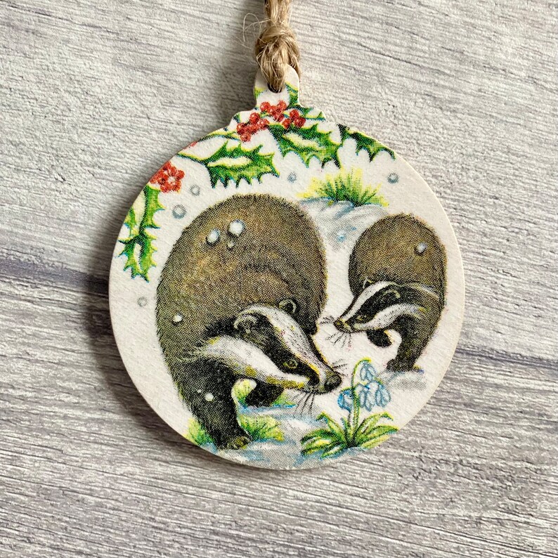 May include: A round wooden ornament featuring a printed illustration of two badgers in a snowy landscape with green holly leaves, red berries, and blue snowdrop flowers. The ornament is suspended by a natural jute twine loop at the top.