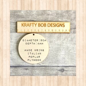 May include: A circular light-colored wooden disc with a small hole at the top. The text on the disc reads: DIAMETER: 9CM, DEPTH: 4MM, MADE USING ITALIAN POPLAR PLYWOOD. A wooden ruler labeled KRAFTY BOB DESIGNS is positioned above the disc for scale.