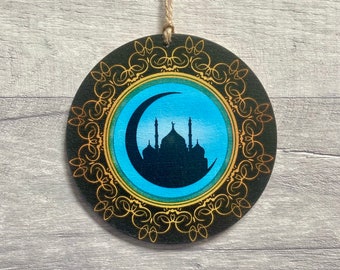 May include: A circular wooden ornament featuring a black silhouette of a mosque and a crescent moon against a bright blue background. The design is framed by an intricate gold filigree border on a black background, suspended by a twine loop.
