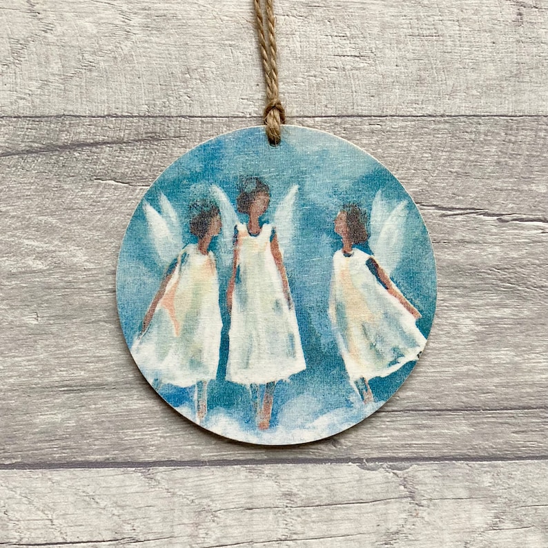 May include: A circular wooden ornament featuring a painted illustration of three winged figures in white dresses against a blue background, suspended by a thin brown twine loop attached to the top center of the round wooden disc.