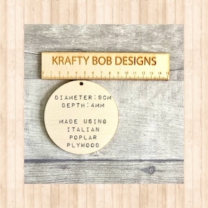 May include: A circular light-colored wooden disc with a small hole at the top. Text on the disc reads: DIAMETER: 9CM, DEPTH: 4MM, MADE USING ITALIAN POPLAR PLYWOOD. A wooden ruler labeled KRAFTY BOB DESIGNS is positioned above the disc.