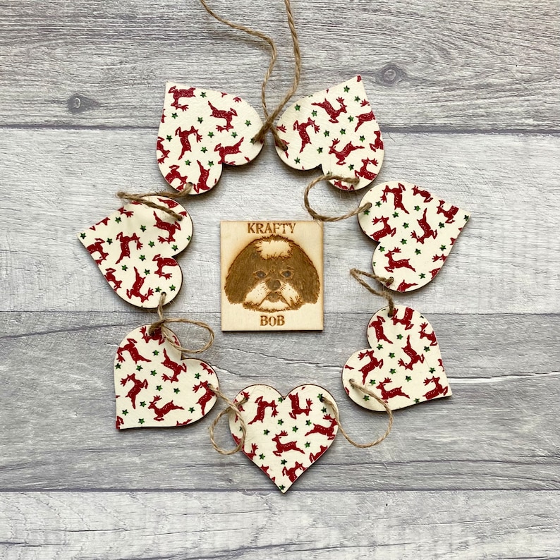 May include: A set of six wooden heart-shaped ornaments featuring a red reindeer and green star pattern, connected by twine. In the center is a square wooden plaque with a wood-burned illustration of a dog and the text KRAFTY BOB.