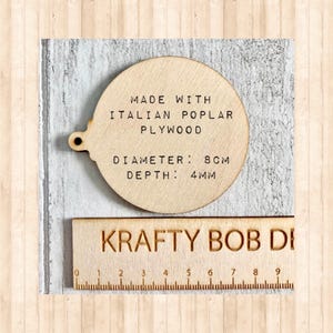 May include: A circular light-coloured wooden ornament with a small hole on the left side. The text reads: MADE WITH ITALIAN POPLAR PLYWOOD, DIAMETER: 8CM, DEPTH: 4MM. A wooden ruler with the text KRAFTY BOB DE is positioned below the ornament.