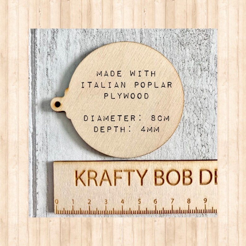 May include: A circular light-coloured wooden ornament with a small hole on the left side. The text reads: MADE WITH ITALIAN POPLAR PLYWOOD, DIAMETER: 8CM, DEPTH: 4MM. A wooden ruler with the text KRAFTY BOB DE is positioned below the ornament.