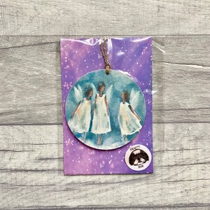 May include: A round wooden ornament featuring three winged figures in white dresses against a blue background, suspended by a jute string. The ornament is packaged in a purple card with white speckles and a circular sticker labeled KRAFTY BOB.