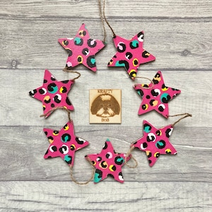 May include: A garland of pink wooden star decorations with a leopard print pattern in black, yellow, and teal. A small wooden square with a dog portrait and the text "KRAFTY BOB" is in the centre.