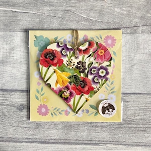 A wooden heart-shaped ornament with a floral pattern. The ornament is decorated with a variety of flowers in shades of red, pink, purple, yellow, and green. The ornament is hanging with natural twine.