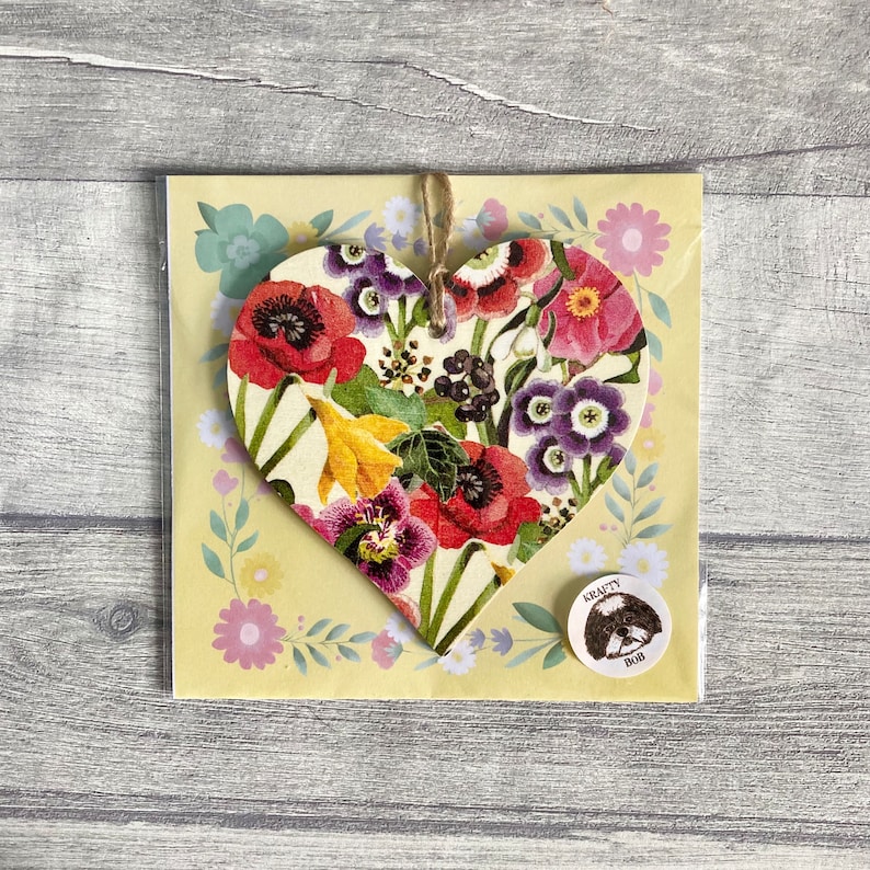 A wooden heart-shaped ornament with a floral pattern. The ornament is decorated with a variety of flowers in shades of red, pink, purple, yellow, and green. The ornament is hanging with natural twine.
