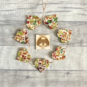 May include: A circular arrangement of six wooden heart-shaped ornaments featuring a colourful autumn leaf pattern, connected by twine. In the centre, a square wooden plaque displays a wood-burned illustration of a dog with the text KRAFTY BOB.
