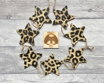 Puede incluir: Una guirnalda de siete adornos en forma de estrella con un estampado de leopardo en negro y beige, unidos por un cordel fino. En el centro, una placa cuadrada de madera muestra un grabado láser de un perro con el texto KRAFTY BOB.
