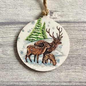 May include: A round wooden ornament featuring a printed illustration of a stag and two deer standing in a snowy landscape with green pine trees. The ornament has a light-coloured wood grain finish and includes a jute twine loop for hanging.