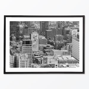 Puede incluir: Una fotograf&iacute;a a&eacute;rea en blanco y negro de un horizonte urbano con una gran valla publicitaria que presenta a un hombre con un sombrero. La valla publicitaria est&aacute; en el lado de un edificio en el medio de la imagen. La ciudad est&aacute; llena de edificios altos y la foto est&aacute; tomada desde un &aacute;ngulo alto.