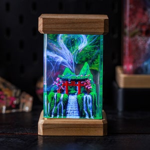 Könnte beinhalten: Eine Holzkiste mit einer Glasfront, die eine Miniatur-Szene mit einem roten japanischen Torii-Tor, einem grünen Berg und einem weißen Wasserfall zeigt. Die Szene wird von hinten beleuchtet, wodurch ein leuchtender Effekt entsteht.