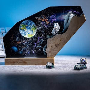 以下が含まれることがあります： 惑星、星、宇宙飛行士が描かれた宇宙のシーンを樹脂で埋め込んだ木製の銘板。銘板は、カラフルな樹脂製の小さなキーキャップが3つ置かれたテーブルの上に置かれている。