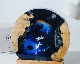 May include: A round resin art piece with a black and blue space scene featuring astronauts floating near a black hole. The resin is set in a wooden base.