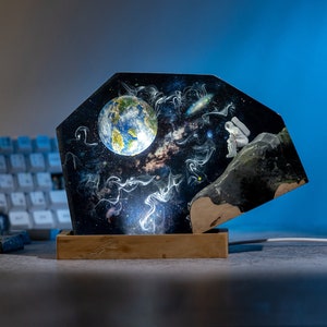 May include: A black and white resin art piece depicting a space scene with a planet, stars, and a figure in a spacesuit. The art piece is mounted on a wooden base.