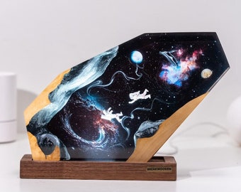 May include: A wooden lamp base with a resin insert featuring a galaxy scene with two astronauts floating in space. The resin is clear and has a black background with a swirling nebula and stars. The base is made of dark wood and has the text "BREAKWOODEN" on the front.