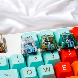 K&ouml;nnte beinhalten: Eine Tastatur mit bunten Tastenkappen mit japanisch inspirierten Designs. Die Tastenkappen zeigen Bilder eines roten Torii-Tores, eines Wasserfalls und eines Waldes. Die Tastatur hat ein hellblaues und wei&szlig;es Farbschema. Die Tasten sind mit Symbolen wie ~, !, @, #, $, %, 1, 2, 3, 4, 5, Q, W, E, R und TAB beschriftet.