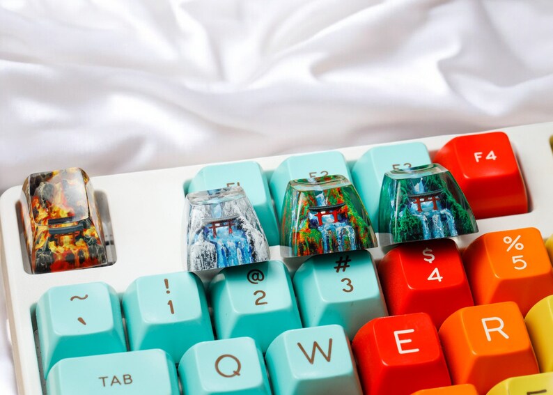 K&ouml;nnte beinhalten: Eine Tastatur mit bunten Tastenkappen mit japanisch inspirierten Designs. Die Tastenkappen zeigen Bilder eines roten Torii-Tores, eines Wasserfalls und eines Waldes. Die Tastatur hat ein hellblaues und wei&szlig;es Farbschema. Die Tasten sind mit Symbolen wie ~, !, @, #, $, %, 1, 2, 3, 4, 5, Q, W, E, R und TAB beschriftet.