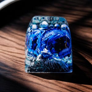 Puede incluir: Un keycap de resina azul con un diseño detallado de un dragón en un patrón de nubes arremolinadas. El keycap es transparente y muestra el dragón con detalle.