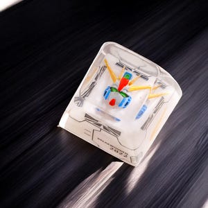 May include: A clear resin cube keycap with a white and yellow robot figure inside. The keycap has the text "BREAKWATER 2022" printed on the bottom.