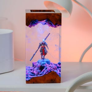 May include: A rectangular wooden and resin lamp featuring a detailed figurine of a warrior holding a staff. The figurine stands on a purple and blue mountain-like structure within the clear resin. The lamp has a warm, ambient glow.