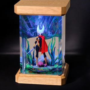Puede incluir: Una lámpara de resina hecha a mano con una figura detallada de un personaje del videojuego Hollow Knight. La figura está representada con una espada y se encuentra sobre una base con detalles de resina verde y azul. La lámpara está iluminada con luces LED azules, creando un efecto mágico y etéreo.