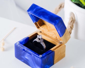 Op de afbeelding: Een blauwe en gouden ringdoos met een zilveren ring erin. De doos is open en onthult een zwart fluwelen interieur. De doos heeft een gemarmerde blauwe hars buitenkant en goudkleurige houten accenten. De ring heeft een grote, heldere edelsteen.