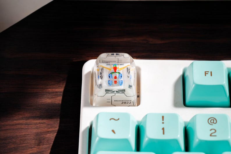 May include: A clear plastic keycap with a white and blue design and the text "DEACTIVATED IN 2022". The keycap is on a white keyboard with turquoise keys labeled "F1", "~", "!", and "@".