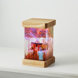 Op de afbeelding: Een houten lampenvoet met een transparante harsinzet met een miniatuur rode tempel en een wervelend roze en oranje wolkachtig ontwerp.