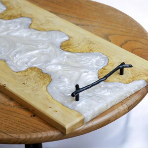 May include: A wooden serving tray with a white resin inlay and black metal handles. The resin has a swirling pattern.