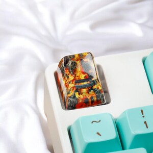 K&ouml;nnte beinhalten: Eine Nahaufnahme eines benutzerdefinierten Tastatur-Keycaps mit einem feurigen orange-schwarzen Design. Der Keycap ist aus Harz gefertigt und hat eine gl&auml;nzende Oberfl&auml;che. Der Keycap befindet sich auf einer wei&szlig;en Tastatur.
