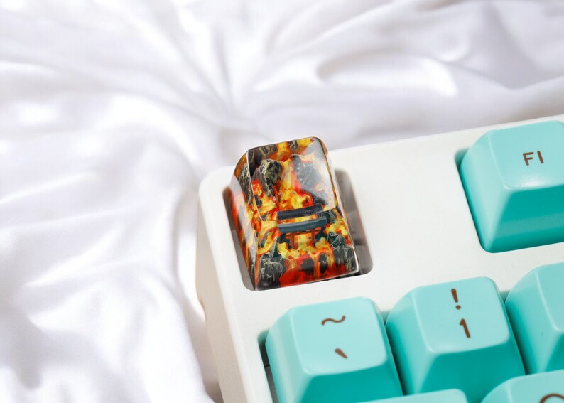 K&ouml;nnte beinhalten: Eine Nahaufnahme eines benutzerdefinierten Tastatur-Keycaps mit einem feurigen orange-schwarzen Design. Der Keycap ist aus Harz gefertigt und hat eine gl&auml;nzende Oberfl&auml;che. Der Keycap befindet sich auf einer wei&szlig;en Tastatur.