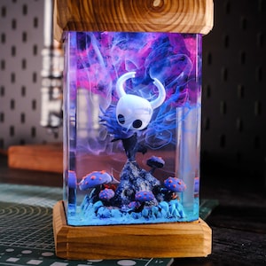 Puede incluir: Una lámpara de resina transparente con una base de madera, que presenta una pequeña figura del Caballero de Hollow Knight, rodeada de remolinos de resina púrpura y azul y decoraciones en miniatura con forma de hongo.