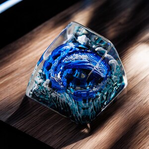 May include: A blue and white resin keycap with a detailed design of a dragon emerging from the water. The keycap is encased in clear resin, giving it a 3D effect.
