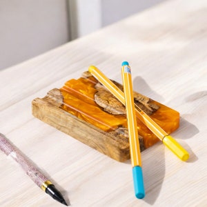 May include: Three yellow pencils with blue tips are placed on a wooden tray with a resin inlay. The tray has a natural wood grain and a yellow and orange resin design.