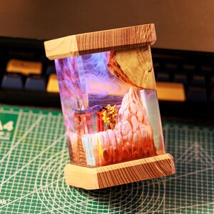 Op de afbeelding: Een houten doos met een transparante harsinzet met een miniatuur sc&egrave;ne van een bergketen met een rode torii-poort en een kleine boom. De hars is gevuld met paarse en blauwe rookachtige swirls.