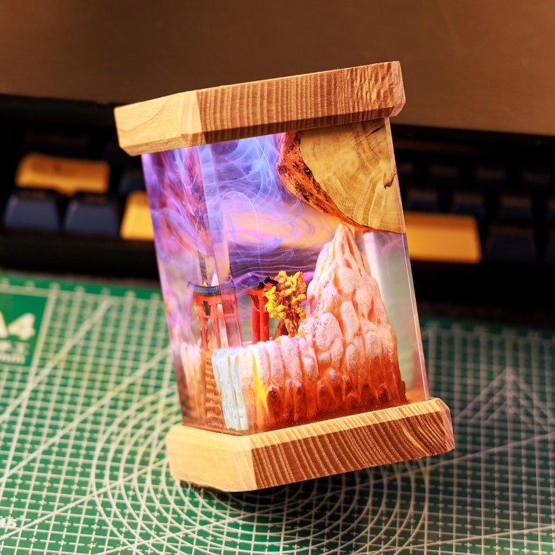 Op de afbeelding: Een houten doos met een transparante harsinzet met een miniatuur sc&egrave;ne van een bergketen met een rode torii-poort en een kleine boom. De hars is gevuld met paarse en blauwe rookachtige swirls.