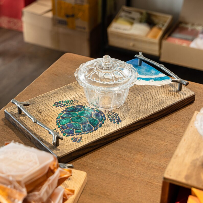 May include: A wooden serving tray with a blue and green sea turtle design. The tray has metal handles shaped like branches. A clear glass jar with a lid is on the tray.