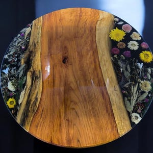 May include: Round wooden table with a glossy finish. The table's center is natural wood, while the edges are embedded with dried flowers and resin. Yellow, white, and purple flowers are visible. The table has a diameter of approximately 30 inches.