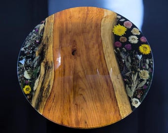 May include: Round wooden table with a glossy finish. The table's center is natural wood, while the edges are embedded with dried flowers and resin. Yellow, white, and purple flowers are visible. The table has a diameter of approximately 30 inches.