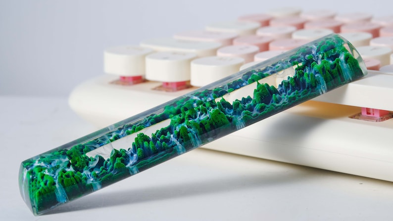 May include: A clear, cylindrical, green and blue resin keycap with a swirling, abstract design. The keycap is resting on a white keyboard.