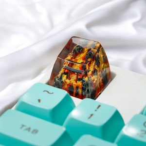 K&ouml;nnte beinhalten: Nahaufnahme eines Tastaturkappen mit einem Harzdesign, das ein feuriges orange-schwarzes Muster zeigt. Die Tastaturkappe befindet sich auf einer wei&szlig;en Tastatur mit blauen Tasten.